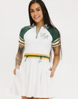 A woman wearing a white dress with a ribbed collar, sleeve cuffs, and a waistband. The dress features a green and yellow stripe detail on the sleeves and a zipper placket, with decorative logos on the pocket.