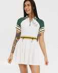 A female model wearing a white dress with a ribbed collar, sleeve cuffs, and a waistband. The dress features a green and yellow trim on the collar, a yellow belt, and white sneakers.