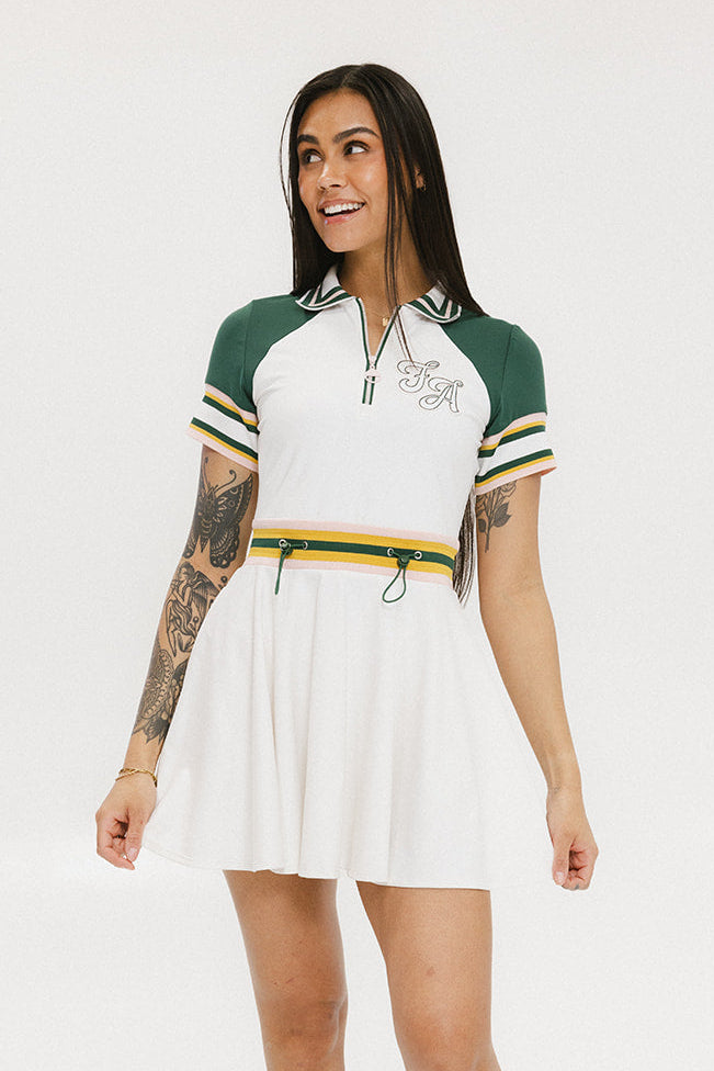 A female model wearing a white dress with a ribbed collar, sleeve cuffs, and a waistband. The dress features a green and yellow trim on the collar, a yellow belt, and white sneakers.