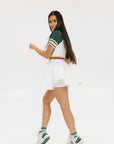 A female model wearing a white dress with a ribbed collar, sleeve cuffs, and a waistband. The dress features a green and yellow trim on the collar, a yellow belt, and white sneakers.