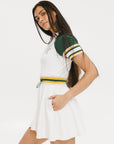 A female model wearing a white dress with a ribbed collar, sleeve cuffs, and a waistband. The dress features a green and yellow trim on the collar, a yellow belt, and white sneakers.