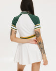 A female model wearing a white dress with a ribbed collar, sleeve cuffs, and a waistband. The dress features a green and yellow trim on the collar, a yellow belt, and white sneakers.