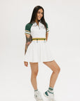 A female model wearing a white dress with a ribbed collar, sleeve cuffs, and a waistband. The dress features a green and yellow trim on the collar, a yellow belt, and white sneakers.