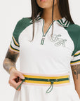 A female model wearing a white dress with a ribbed collar, sleeve cuffs, and a waistband. The dress features a green and yellow trim on the collar, a yellow belt, and white sneakers.