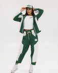 A pair of color-blocked green and white warm-up pants with a pink stripe, featuring a flat drawstring waistband and deep front pockets, branded with the 'Fore All' detail on the back and an FA icon on the inner calf.