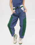 A pair of indigo and green color-blocked warm-up pants with a flat drawstring waistband, deep front pockets, and a barrel style fit. The pants feature appliqué logos, including the 'Fore All' detail on the back and an FA icon on the inner calf.