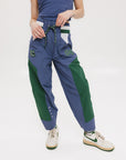 A pair of indigo and green color-blocked warm-up pants with a flat drawstring waistband, deep front pockets, and a barrel style fit. The pants feature appliqué logos, including the 'Fore All' detail on the back and an FA icon on the inner calf.