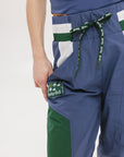 A pair of indigo and green color-blocked warm-up pants with a flat drawstring waistband, deep front pockets, and a barrel style fit. The pants feature appliqué logos, including the 'Fore All' detail on the back and an FA icon on the inner calf.