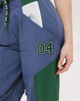 A pair of indigo and green color-blocked warm-up pants with a flat drawstring waistband, deep front pockets, and a barrel style fit. The pants feature appliqué logos, including the 'Fore All' detail on the back and an FA icon on the inner calf.