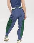 A pair of indigo and green color-blocked warm-up pants with a flat drawstring waistband, deep front pockets, and a barrel style fit. The pants feature appliqué logos, including the 'Fore All' detail on the back and an FA icon on the inner calf.