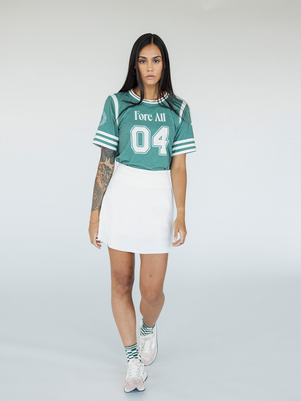A woman wearing a green sporty top with text 'Fore All 04' and stripes, paired with a white skirt.