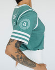 A woman wearing a green sporty top with text 'Fore All 04' and stripes, paired with a white skirt.