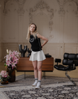 A person standing in a room with classic decor, wearing an off-white knee-length skirt with a black top, accessorized with a necklace and earrings.
