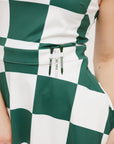 A woman wearing a green and white checkered sleeveless dress with a button placket and side seam pockets.