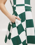 A woman wearing a green and white checkered sleeveless dress with a button placket and side seam pockets.