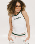 A female model wearing a white sleeveless tank top with a green and pink stripe at the neckline and armholes, and the text 'Chasing Balls' on the chest. She is also wearing white pants with pink and green accents and sunglasses.