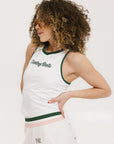 A female model wearing a white sleeveless tank top with a green and pink stripe at the neckline and armholes, and the text 'Chasing Balls' on the chest. She is also wearing white pants with pink and green accents and sunglasses.