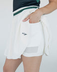 A woman wearing a white pleated golf skirt with a dark color waistband, paired with a gray top and white sneakers.