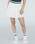 A woman wearing a white pleated golf skirt with a dark color waistband, paired with a gray top and white sneakers.