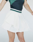 A woman wearing a white pleated golf skirt with a dark color waistband, paired with a gray top and white sneakers.