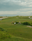 A wide outdoor golf course scene with green fairways, sand bunkers, and a few small people standing near a cart on the course under a cloudy sky.