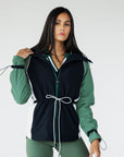 A female model wearing a black rain jacket with green sleeves, featuring a high neck, water-resistant properties, and a double zipper closure.
