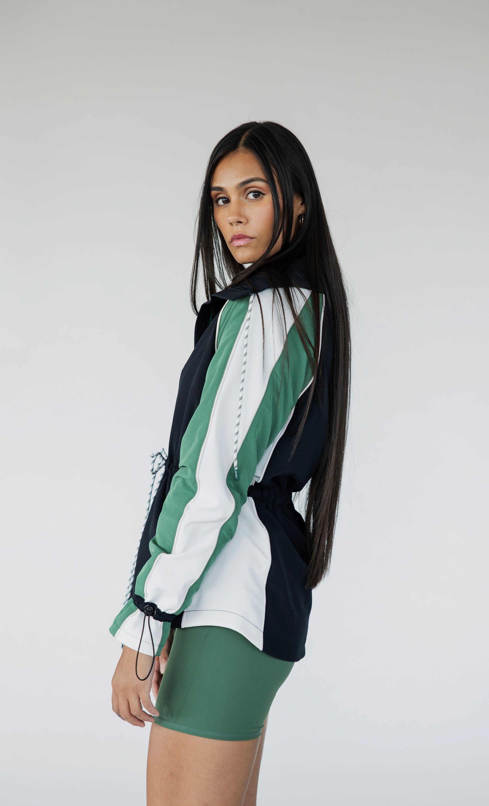 A female model wearing a black rain jacket with green sleeves, featuring a high neck, water-resistant properties, and a double zipper closure.