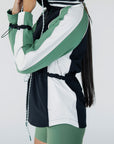 A female model wearing a black rain jacket with green sleeves, featuring a high neck, water-resistant properties, and a double zipper closure.