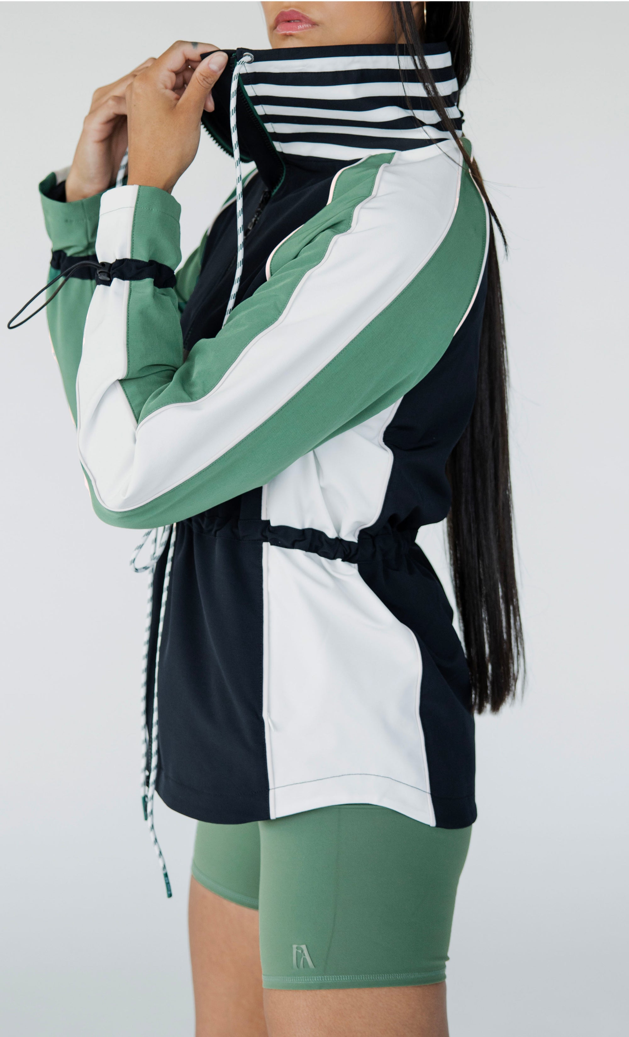 A female model wearing a black rain jacket with green sleeves, featuring a high neck, water-resistant properties, and a double zipper closure.