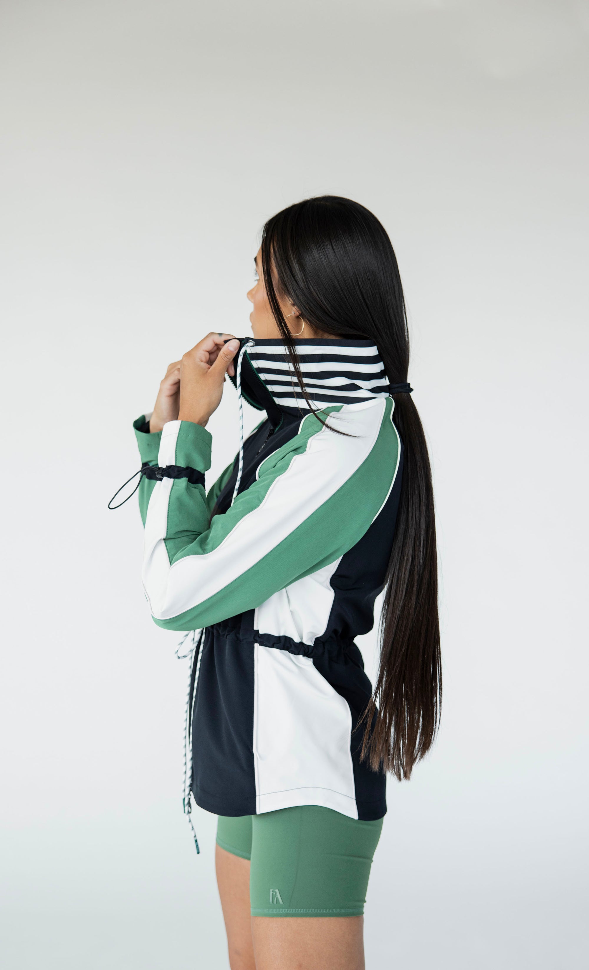 A female model wearing a black rain jacket with green sleeves, featuring a high neck, water-resistant properties, and a double zipper closure.