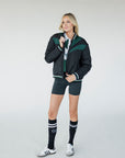 A female model wearing a black bomber jacket with color-blocked sunset design on the back, standing against a neutral background.