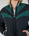A female model wearing a black bomber jacket with color-blocked sunset design on the back, standing against a neutral background.