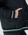 A female model wearing a black bomber jacket with color-blocked sunset design on the back, standing against a neutral background.