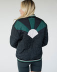A female model wearing a black bomber jacket with color-blocked sunset design on the back, standing against a neutral background.