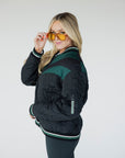 A female model wearing a black bomber jacket with color-blocked sunset design on the back, standing against a neutral background.
