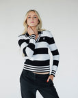 A woman wearing a black and white striped sweater with a V-neck collar and a small embroidered patch on the left chest.