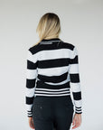 A woman wearing a black and white striped sweater with a V-neck collar and a small embroidered patch on the left chest.