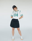 A person wearing a white golf shirt with green and black stripes, featuring a flamingo graphic and the text 'Fore All 04'. They are also wearing a black skirt and a black hat with text.