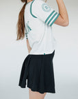 A person wearing a white golf shirt with green and black stripes, featuring a flamingo graphic and the text 'Fore All 04'. They are also wearing a black skirt.