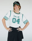 A person wearing a white golf shirt with green and black stripes, featuring a flamingo graphic and the text 'Fore All 04'. They are also wearing a black skirt and a black hat with text.
