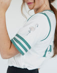 A person wearing a white golf shirt with green and black stripes, featuring a flamingo graphic and the text 'Fore All 04'. They are also wearing a black skirt and a black hat with text.