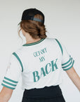 A person wearing a white golf shirt with green and black stripes, featuring a flamingo graphic and the text 'Fore All 04'. They are also wearing a black skirt and a black hat with text.