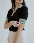 A woman wearing a black polo shirt with a geometric design on the collar and sleeves, and light pink pants.