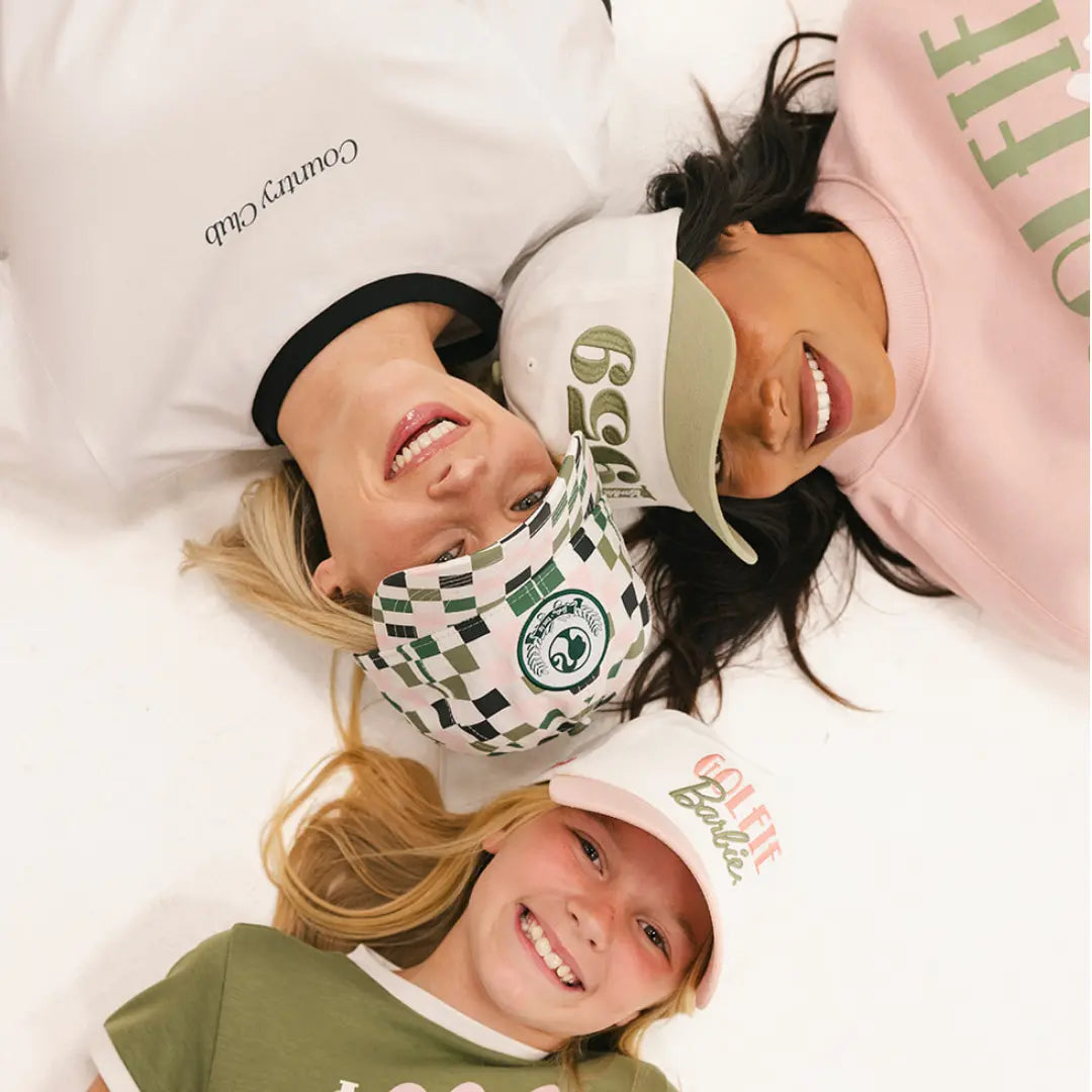 Three smiling golfers lying in a circle wearing Fore All golf hats and Barbie X Fore All golf apparel in soft pink, white, and green tones.