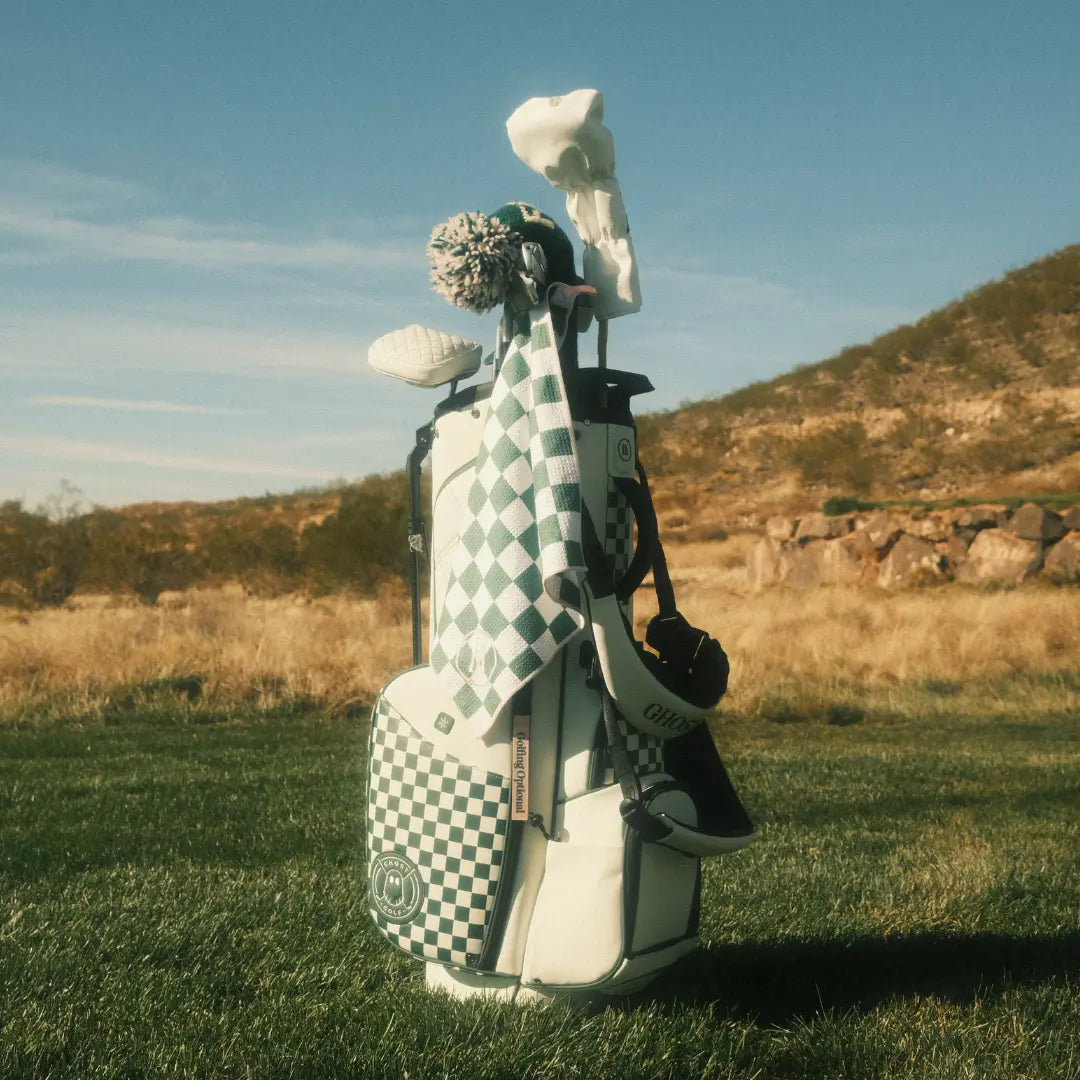 White and green checkered golf bag with matching accessories and club covers on a sunny golf course with desert hills in the background.