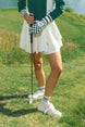 A woman wearing a white pleated golf skirt with a dark color waistband, paired with a green top and pink sneakers.