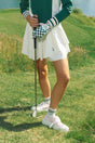 A woman wearing a white pleated golf skirt with a dark color waistband, paired with a green top and pink sneakers.