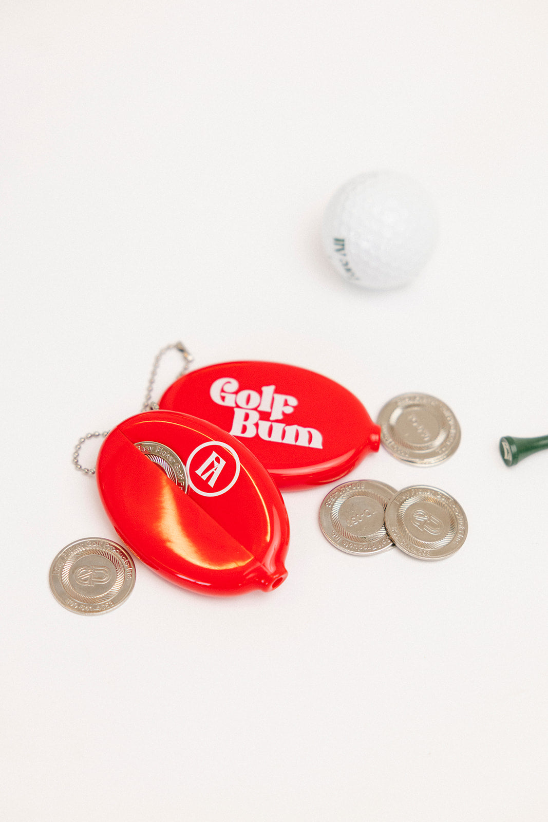 Red 'Golf Bum' golf ball marker with coins and a golf ball on a white background