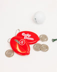 Red 'Golf Bum' golf ball marker with coins and a golf ball on a white background