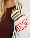 Close-up of a person wearing a white knit sweater with red and black patterns, held against a blurred background.